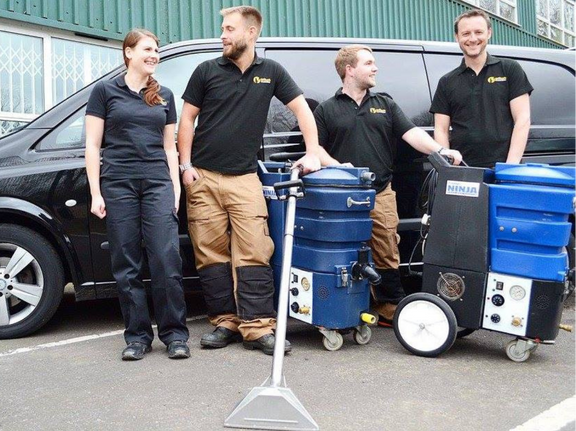 This is a photo of Tooting Carpet Cleaning carpet cleaners (three men and one woman) standing in fromt of their black van, with two steam cleaning carpet machines next to them